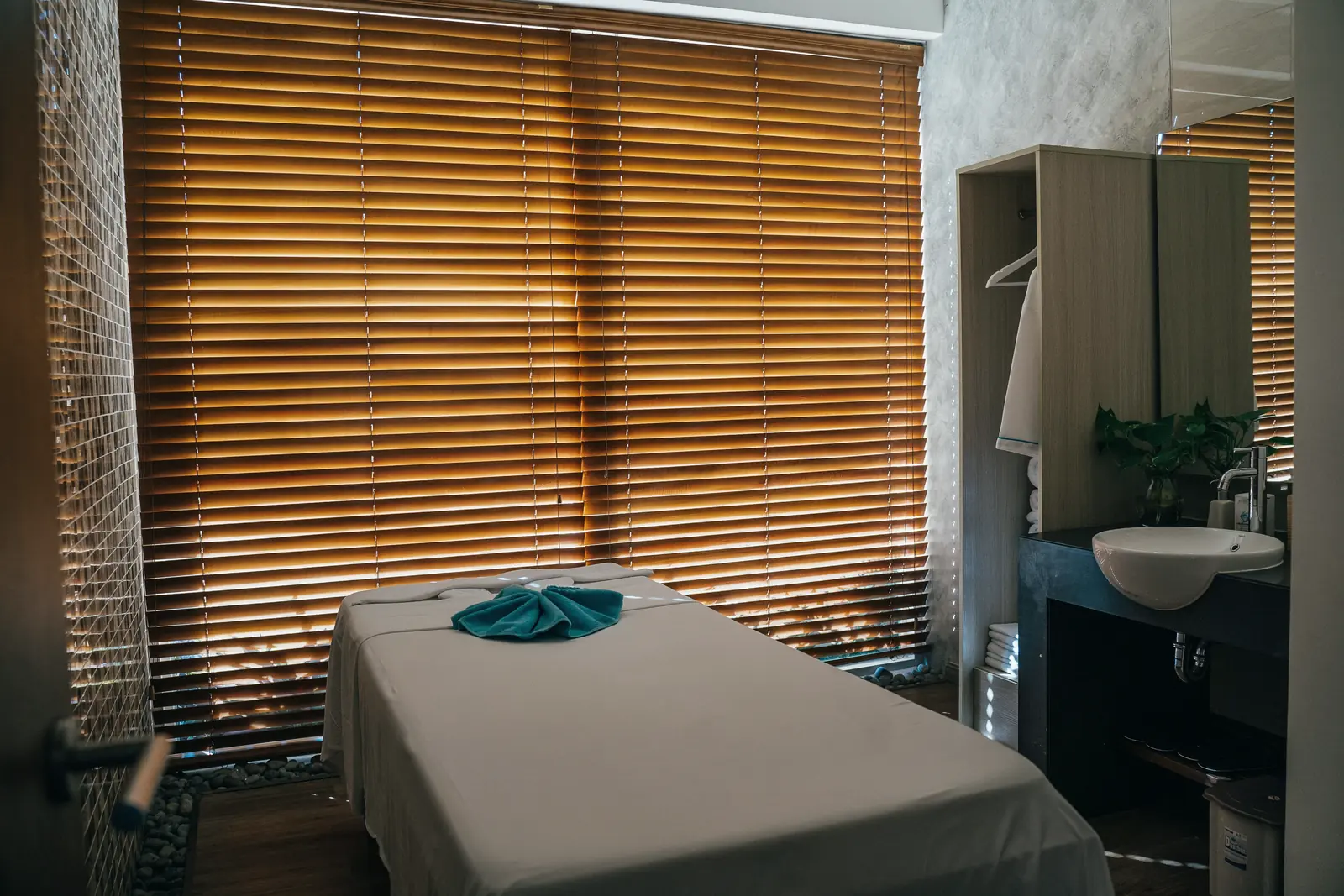 Leere Massageliege mit Handtuch vor einem Fenster mit Lamellenjalousie.