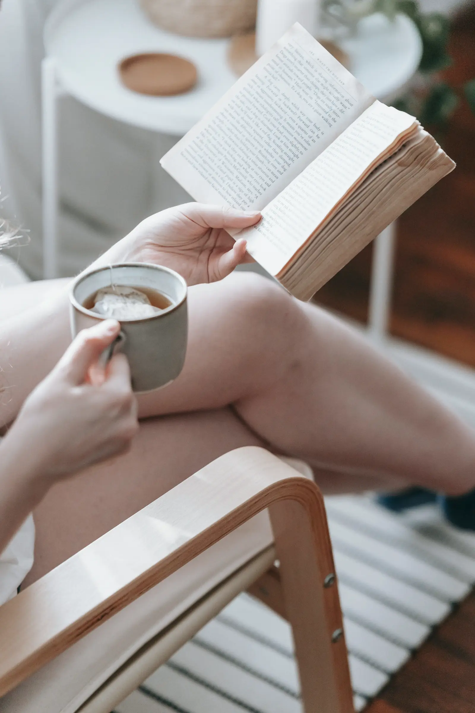 Frau sitzt entspannt zuhause auf dem Sofa, liest ein Buch und trinkt Tee.