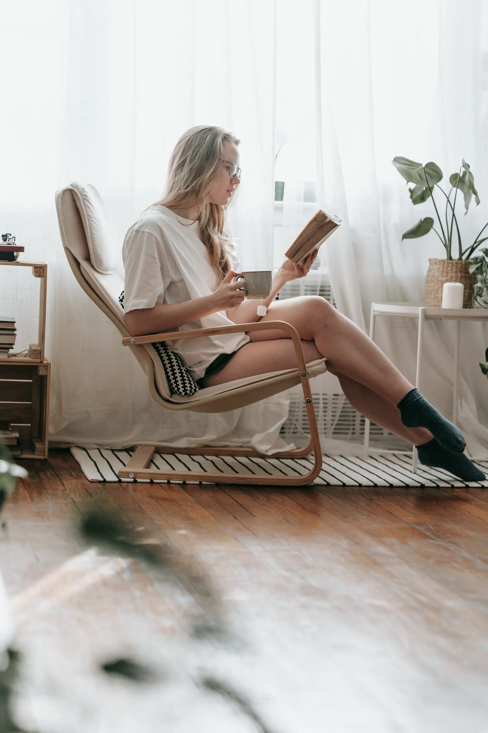 Junge Frau mit Brille liest ein Buch in einem Sessel am Fenster.